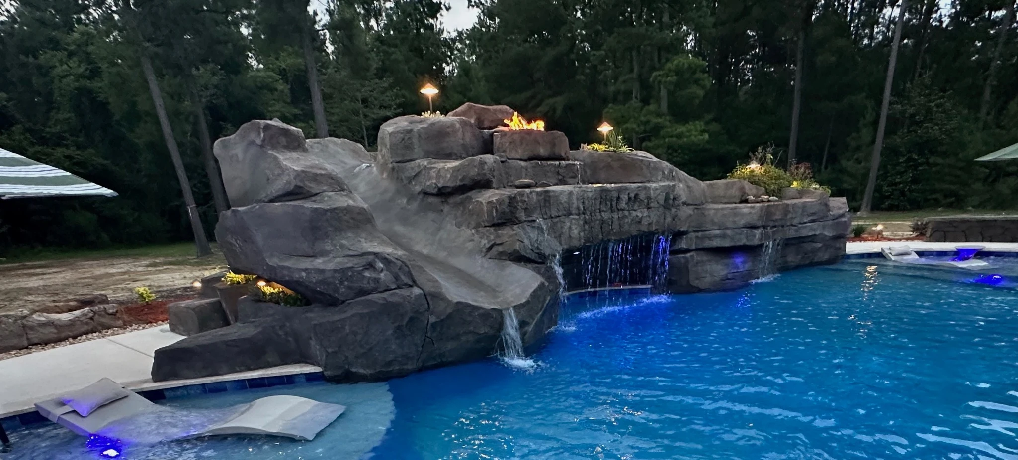 Rock waterfall and grotto with a slide, blue pool lighting and an integrated fire feature at dusk